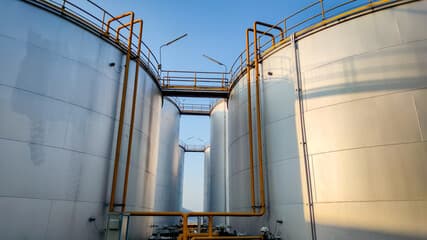 Rows of industrial oil storage tanks at dusk under an amber sky, gauges showing low levels.